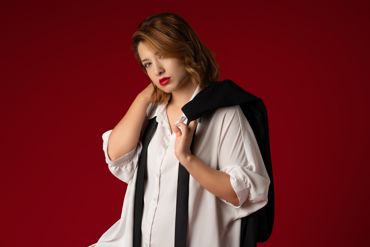 Woman in a white shirt with a black blazer draped over her shoulders against a deep red backdrop. Overworked & Underpaid, a shoot by Nissi Pululu of Sonset Inc in Toronto