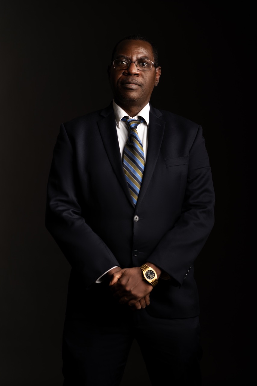 Man in a navy suit and striped tie standing with his hands together against a dark backdrop. Headshots, a shoot by Nissi Pululu of Sonset Inc in Toronto