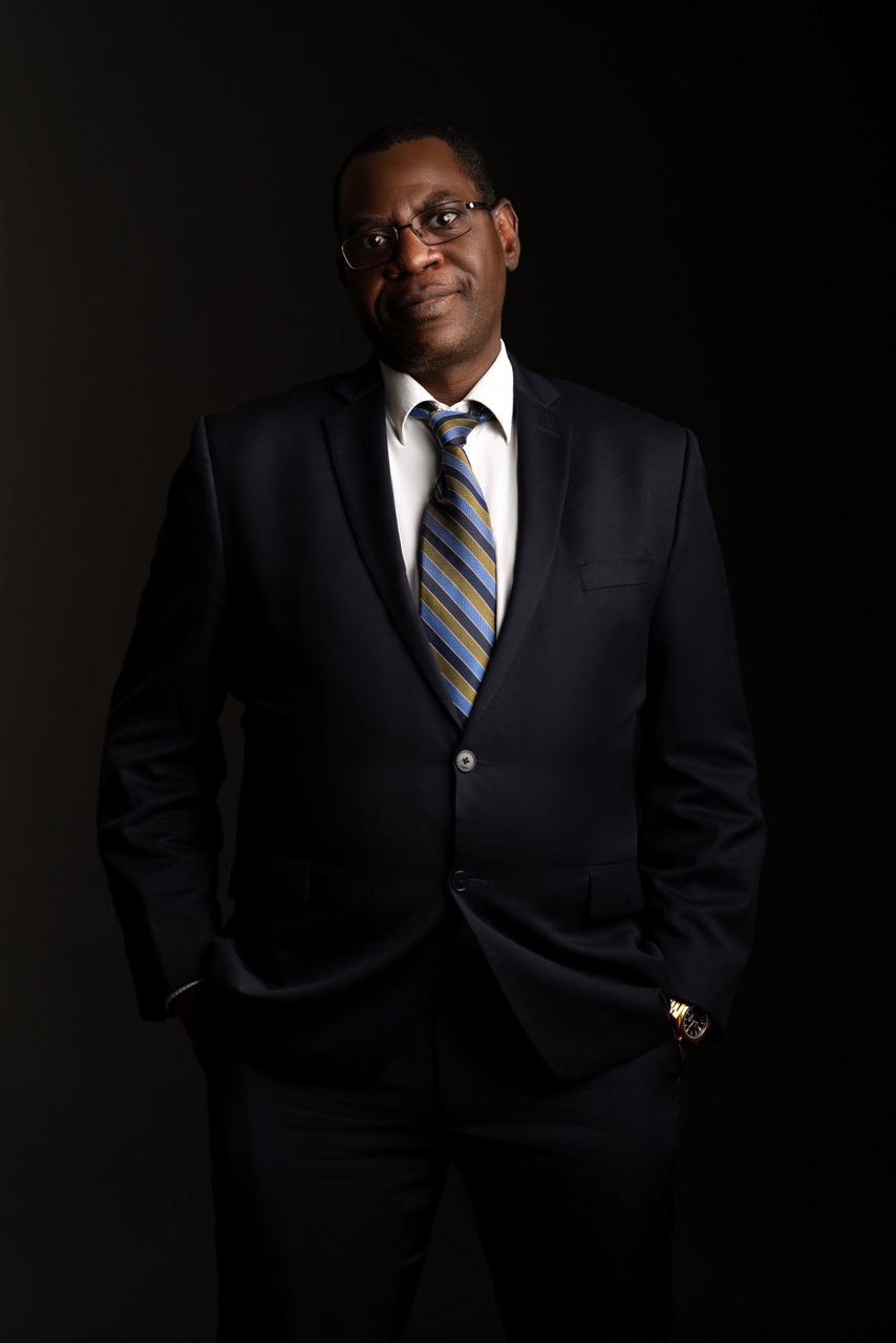 Man in a navy suit and striped tie standing with one hand in his pocket against a dark backdrop. Headshots, a shoot by Nissi Pululu of Sonset Inc in Toronto