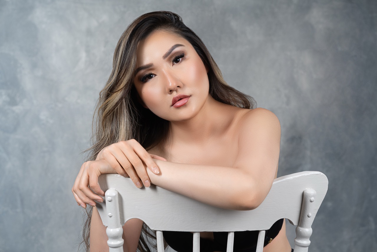 Woman with long light brown hair sitting on a white chair against a grey painted backdrop. Sit For Who?, a shoot by Nissi Pululu of Sonset Inc in Toronto