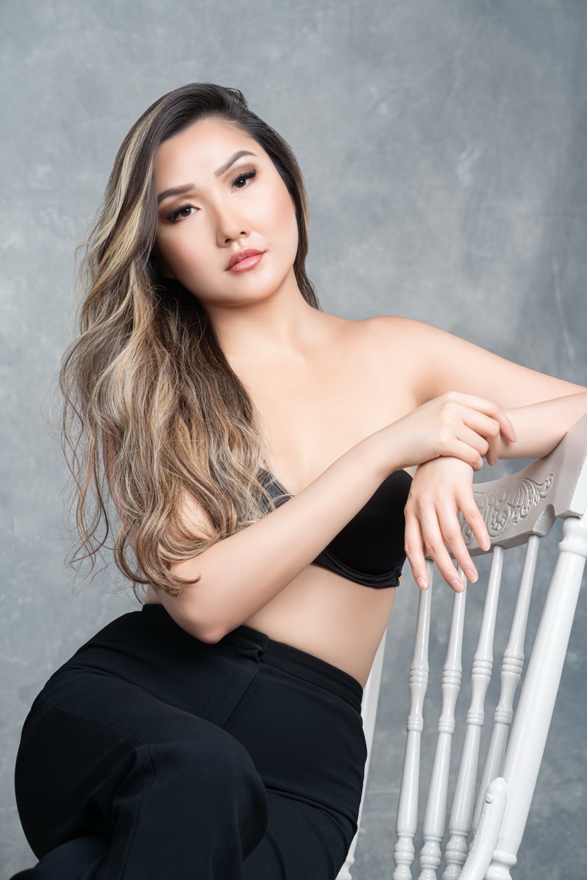 Woman with long wavy hair in a black top and trousers seated on a white chair against a grey painted backdrop. Sit For Who?, a shoot by Nissi Pululu of Sonset Inc in Toronto