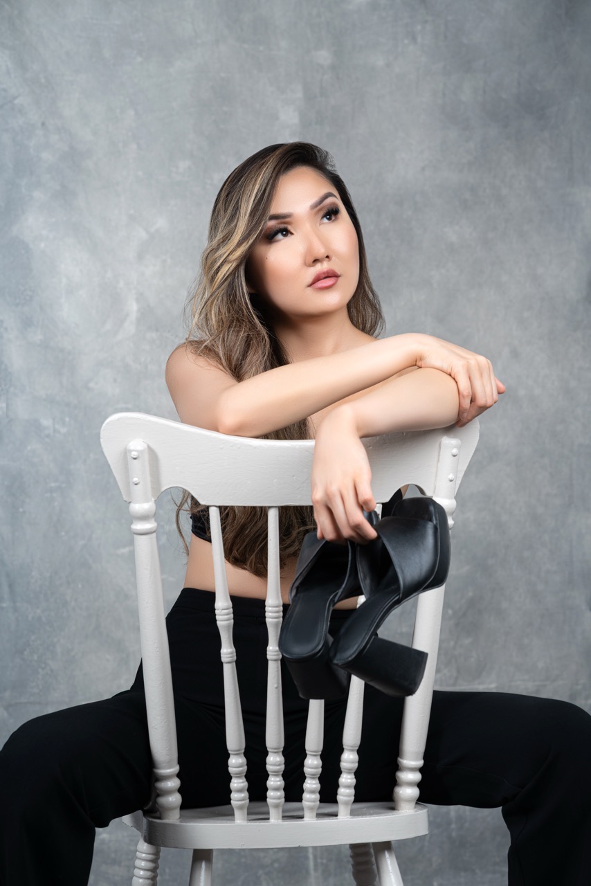 Woman in black trousers sitting backwards on a white chair holding black platform shoes against a grey painted backdrop. Sit For Who?, a shoot by Nissi Pululu of Sonset Inc in Toronto