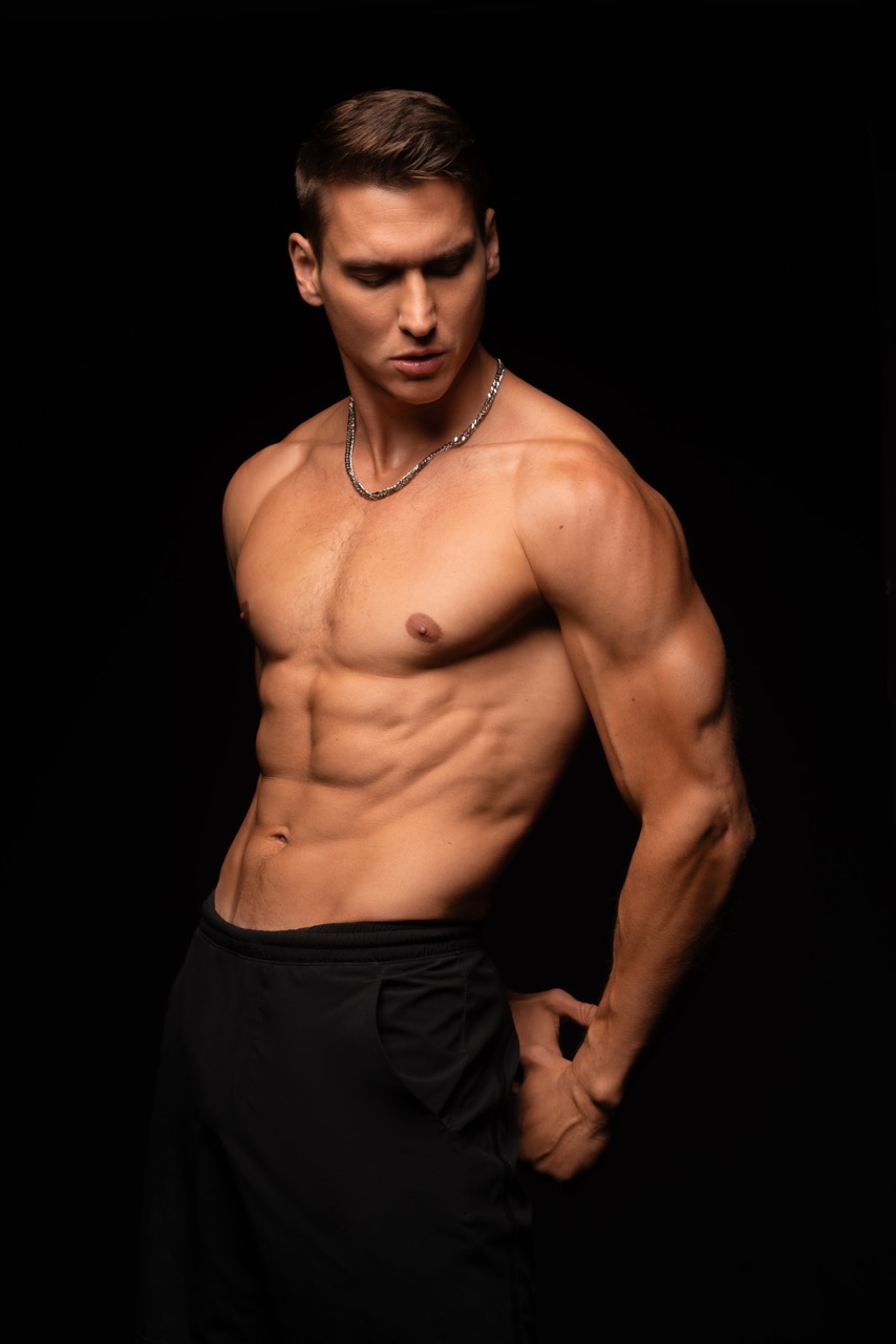 Shirtless muscular man in black trousers looking down against a black backdrop. Mind Vs Body, a shoot by Nissi Pululu of Sonset Inc in Toronto