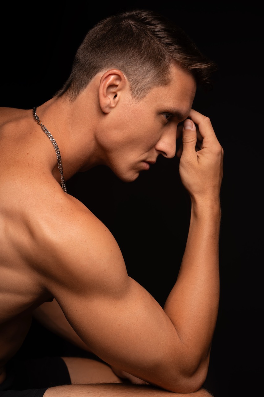 Shirtless muscular man with his hand on his forehead, looking down against a dark backdrop. Mind Vs Body, a shoot by Nissi Pululu of Sonset Inc in Toronto