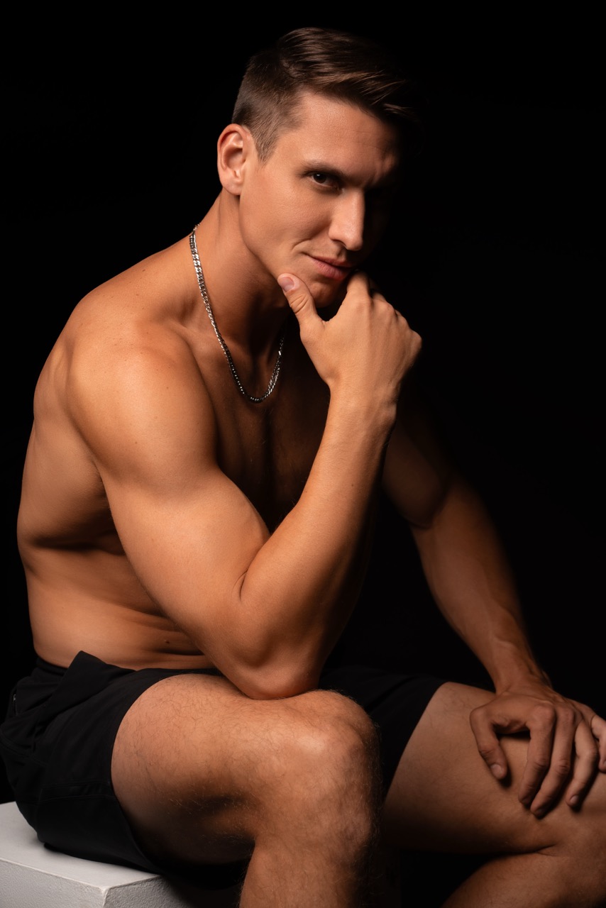Shirtless muscular man in black shorts sitting on a white block with his hand at his chin against a black backdrop. Mind Vs Body, a shoot by Nissi Pululu of Sonset Inc in Toronto