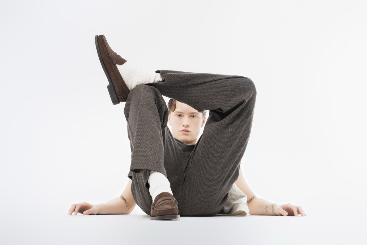 Man in grey trousers and brown shoes lying on a white floor with his legs in the air against a white backdrop. Titanic, a shoot by Nissi Pululu of Sonset Inc in Toronto