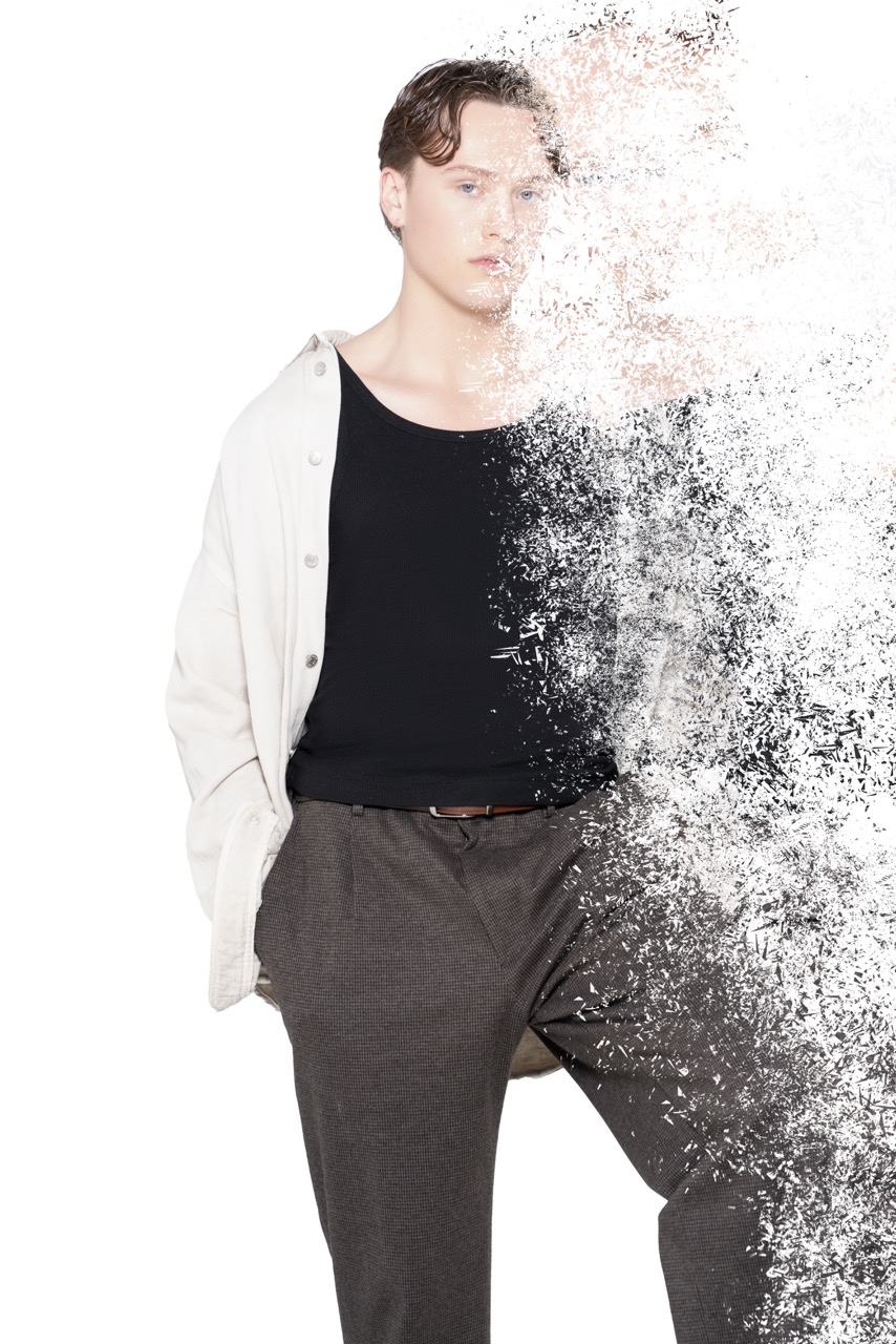 Man in a black top and grey trousers with half of him dissolving into particles against a white backdrop. Titanic, a shoot by Nissi Pululu of Sonset Inc in Toronto