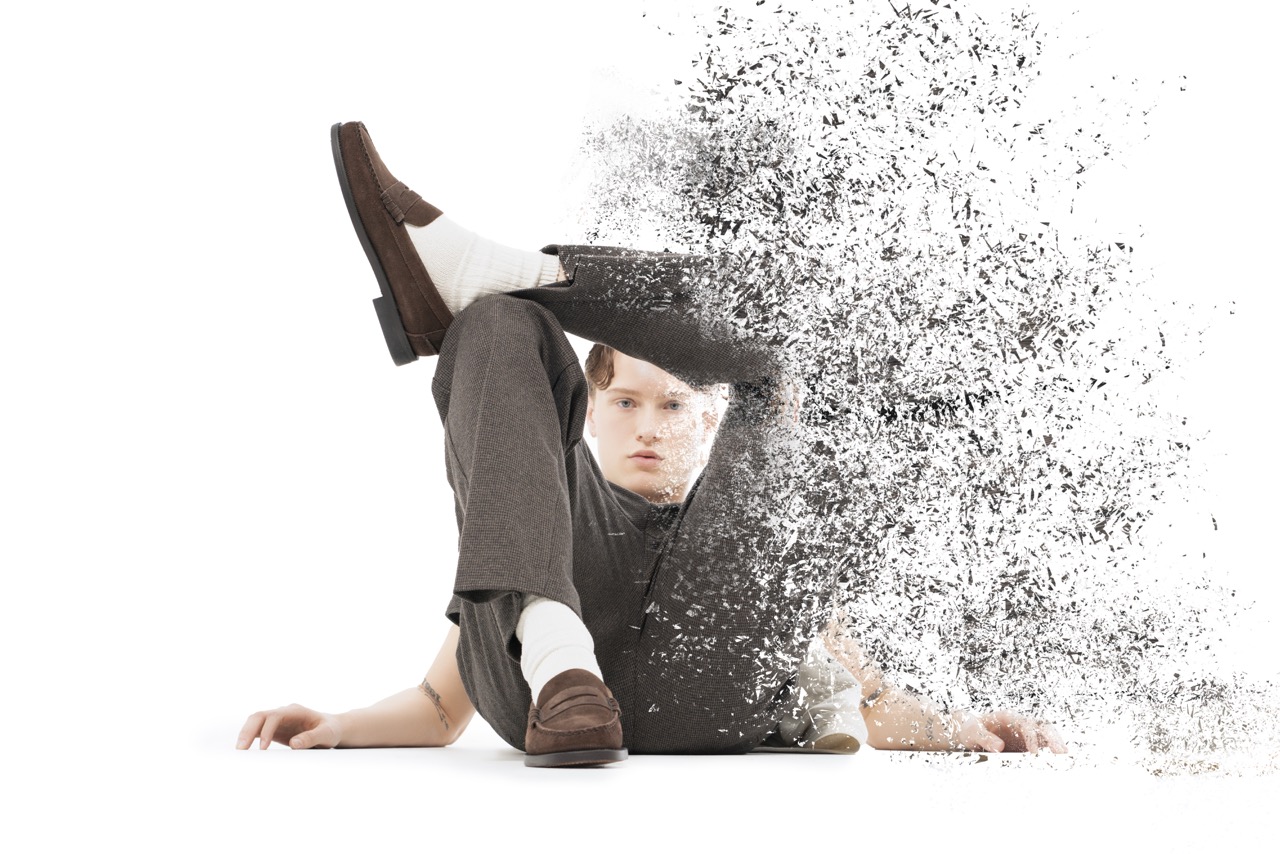Man in grey trousers lying on a white floor with his body dissolving into particles. Titanic, a shoot by Nissi Pululu of Sonset Inc in Toronto