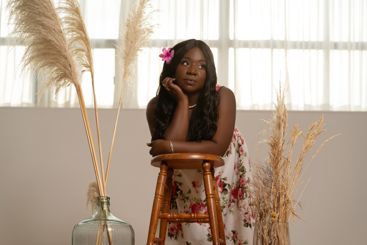 Woman in a floral dress with a flower in her hair leaning on a wooden stool beside tall dried grass. Summertime, a shoot by Nissi Pululu of Sonset Inc in Toronto