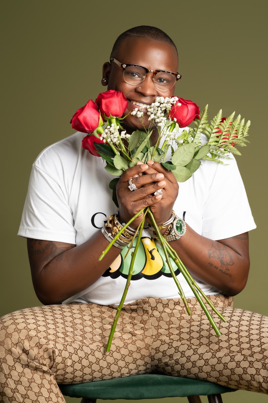 Man in glasses and a white shirt smiling behind a bouquet of red roses against an olive green backdrop. Picture Day, a shoot by Nissi Pululu of Sonset Inc in Toronto