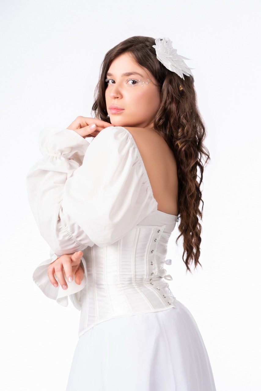 Woman with long dark hair and a white bow in a white corset dress looking over her shoulder against a white backdrop. Angels, a shoot by Nissi Pululu of Sonset Inc in Toronto