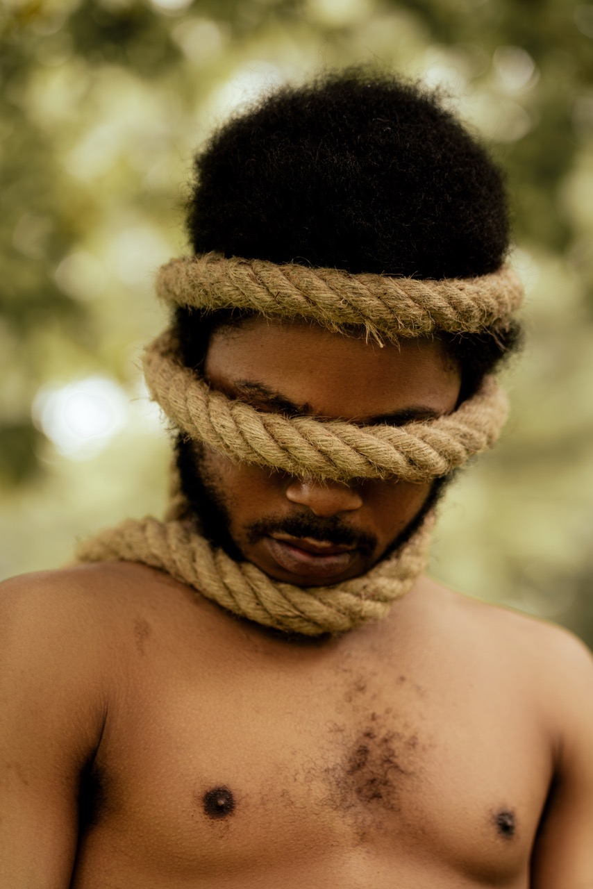 Man with thick rope wrapped around his eyes and neck against a soft green outdoor background. Freedom, a shoot by Nissi Pululu of Sonset Inc in Toronto